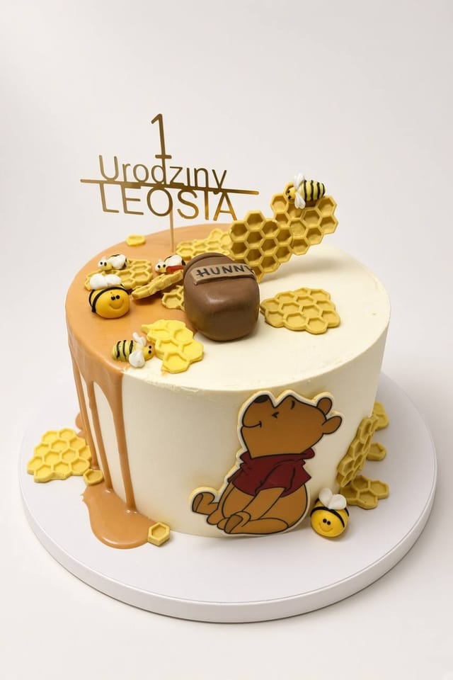 Jasny tort na roczek Leosia z Kubusiem Puchatkiem, pszczółkami, żółtymi plastrami miodu, garnuszkiem HUNNY i złotym topperem „1 Urodziny LEOSIA”.