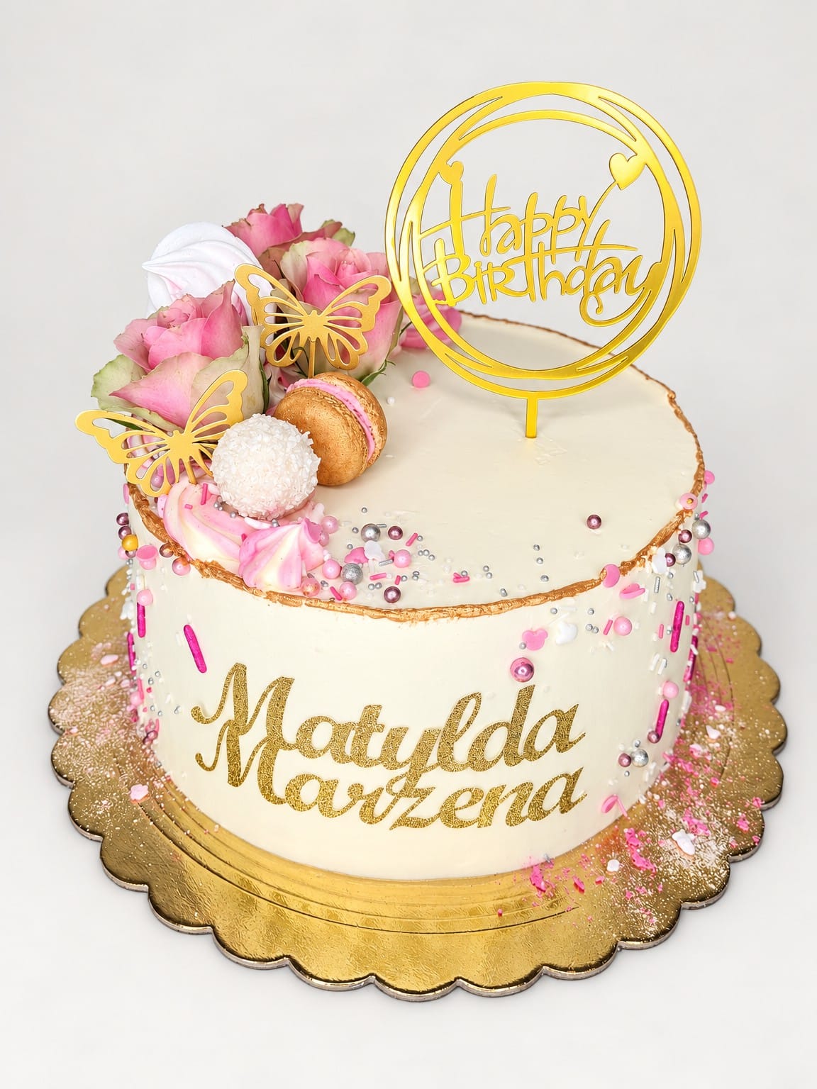 Jasny okrągły tort z różowymi różami, złotymi motylami, topperem „Happy Birthday” i złotymi napisami „Matylda” oraz „Marzena”.