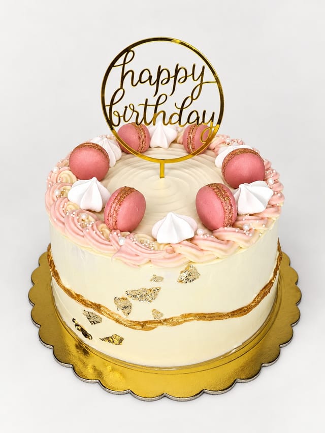 Okrągły jasny tort z różowymi dekoracjami, białymi ozdobami, perełkami i złotym topperem „happy birthday”.