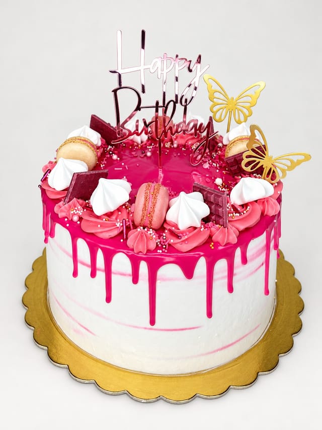 Okrągły biało-różowy tort z różowymi zaciekami, rozetkami, drobnymi ozdobami, dwoma złotymi motylami i napisem „Happy Birthday”.