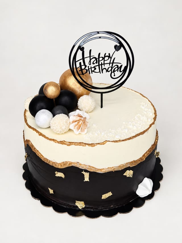 Czarno-biały tort ze złotymi krawędziami, czarnymi, złotymi, srebrnymi i białymi kulami oraz topperem z napisem happy Birthday.