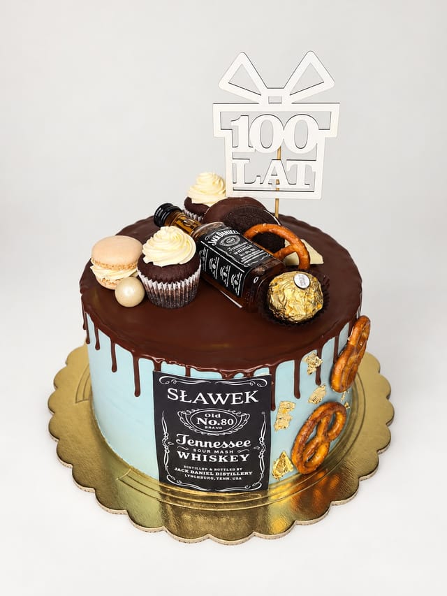 Jasnoniebieski tort z brązowymi zaciekami, etykietą „SŁAWEK”, topperem „100 LAT”, miniaturową butelką, babeczkami i preclami.